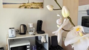 Una habitación con una mesa de comedor, una cafetera, un microondas, un cubo de basura y una fotografía de una playa en la pared.