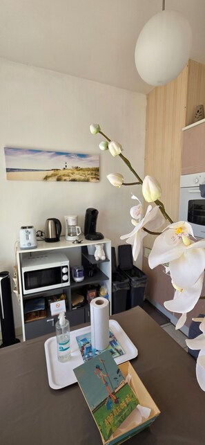 A room with a dining table, a coffee maker, a microwave, a trash bin, and a picture of a beach on the wall.