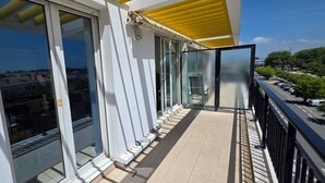 Un balcón con puertas de vidrio, un toldo amarillo y vistas a una calle con coches aparcados y árboles.