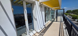 A balcony with glass doors, a yellow awning, and a view of a street with parked cars and trees.