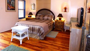 Une chambre à coucher comprenant un lit, deux tables de chevet avec des lampes, un téléviseur, une chaise, une petite table et des œuvres d’art accrochées aux murs.