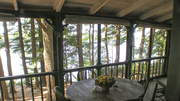 Mesa de madera con sillas en un espacio exterior cubierto, con ventanales grandes y rodeado de árboles.