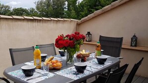 Una mesa de comedor al aire libre con sillas, un jarrón de flores rojas y diferentes alimentos.