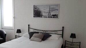 Una cama bien hecha con edredón blanco, cabecero de metal negro y dos lámparas de noche. Encima de la cama cuelga un cuadro con la Torre Eiffel.