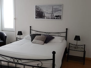 Un lit bien fait, avec une couette blanche, une tête de lit en métal noir et deux lampes de chevet. Un tableau encadré représentant la Tour Eiffel est accroché au-dessus du lit.