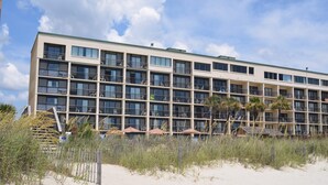 A multi-story hotel with balconies, located near a sandy beach with some vegetation.