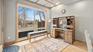 A room with a large window, a desk with a lamp, a bench, and a patterned rug.