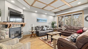 A cozy living room with a fireplace, leather sofas, a flat-screen TV, and a large window offering a view of trees.