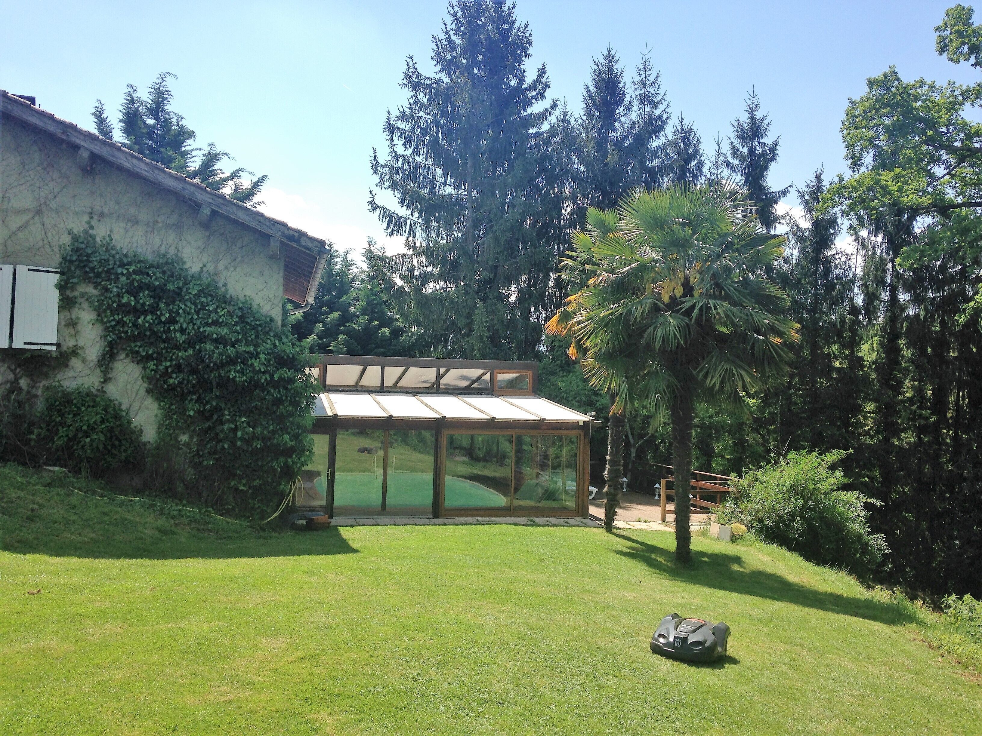 Un jardin avec une terrasse fermée par des baies vitrées, une pelouse et des arbres tout autour.