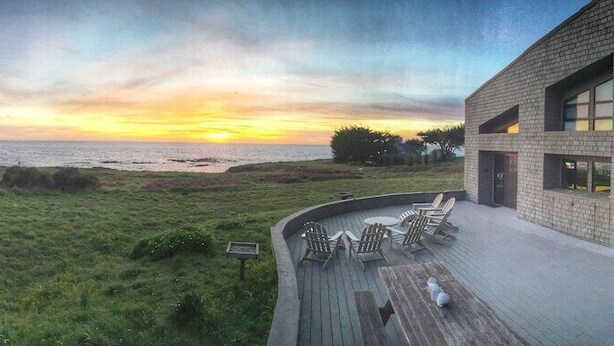 Um edifício moderno com fachada de tijolos, um deck com área de estar externa e vista para o oceano ao pôr do sol.