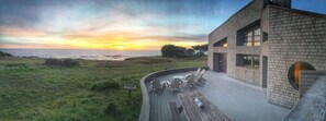 A modern building with a brick facade, a deck with outdoor seating, and a view of the ocean at sunset. - Stunning Oceanfront Home w/ Incredible Ocean Views (The Sea Ranch)