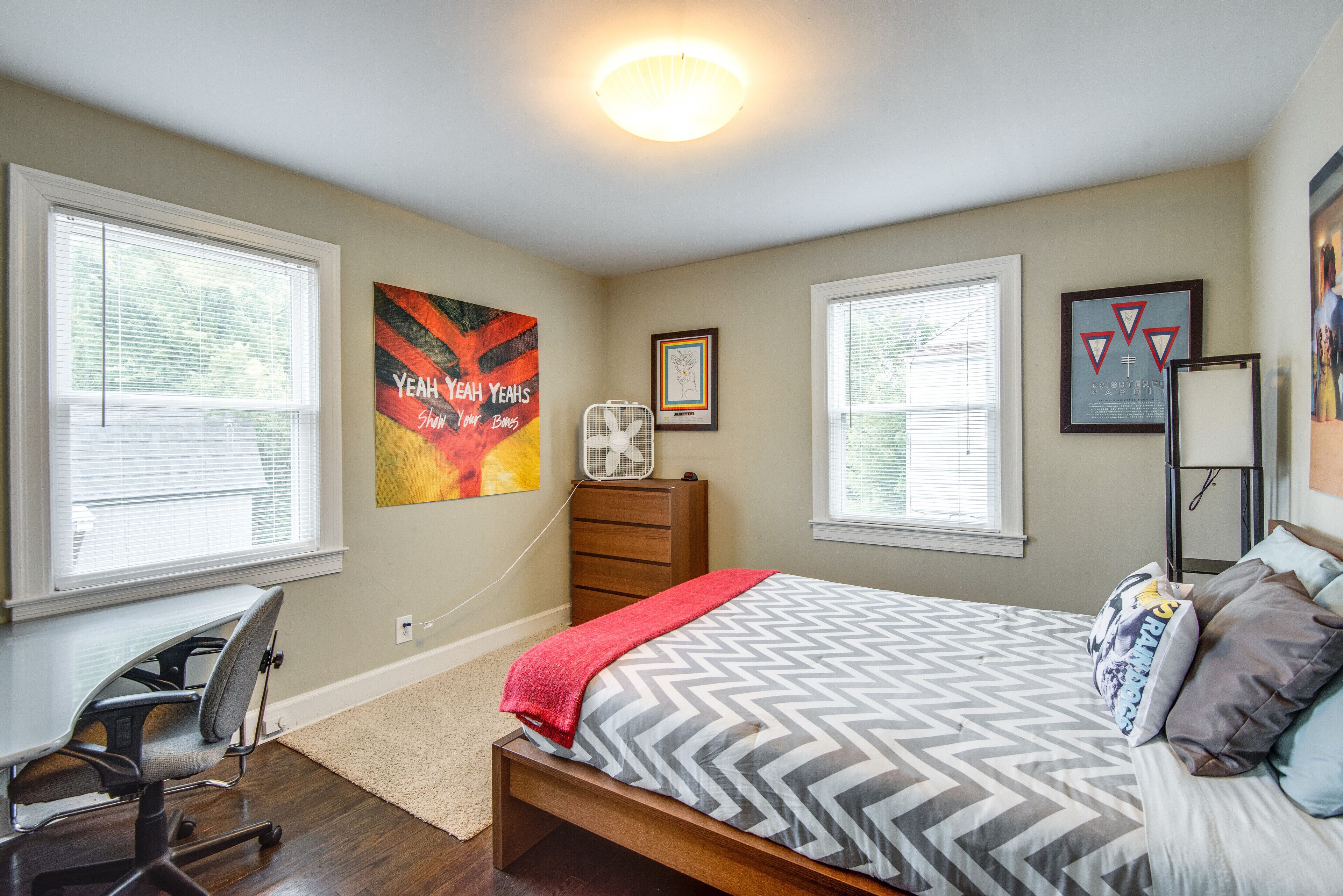 A bedroom with a bed, a desk, and a chair. There are two windows with blinds, a ceiling fan, and wall art.