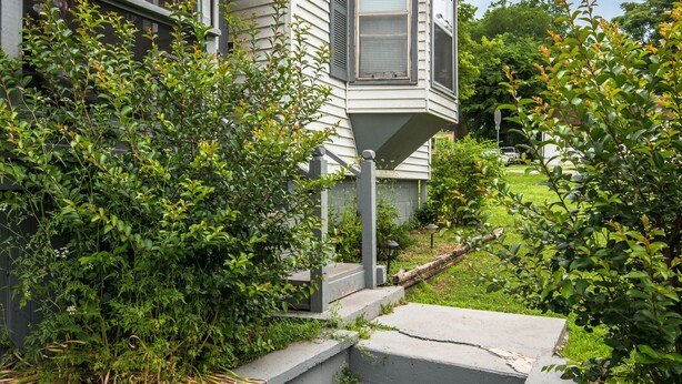 Una casa con un porche, escalones que llevan a la entrada y un jardín con arbustos y plantas.