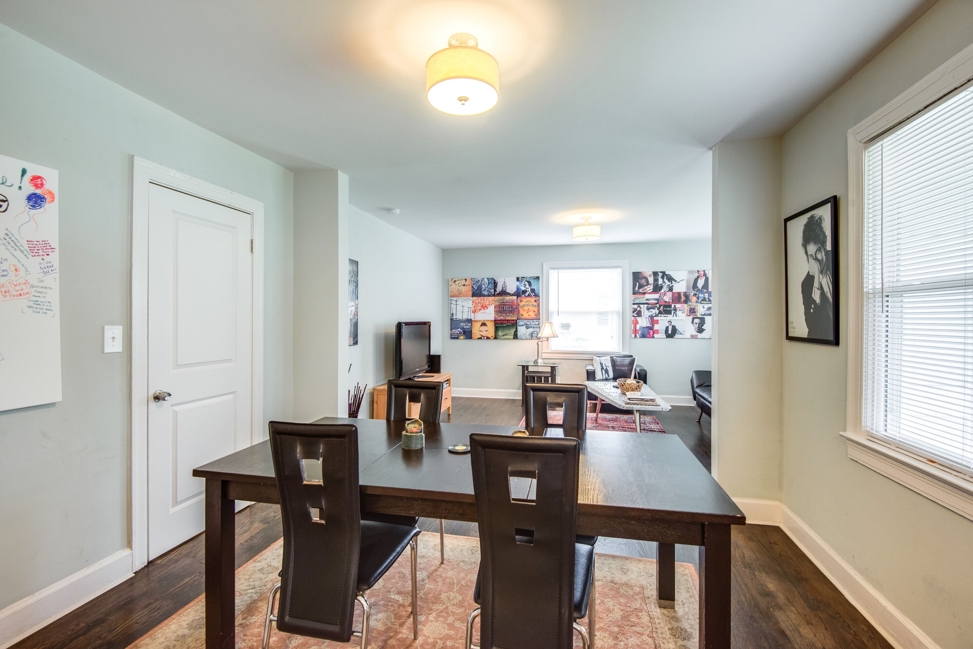 A dining area with a long table, chairs, a television, and a kitchen area in the background.