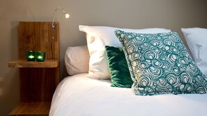 Una cama bien tendida con sábanas blancas y dos almohadas, una verde y otra estampada, en una habitación con una mesita de noche de madera y una taza verde pequeña encima.