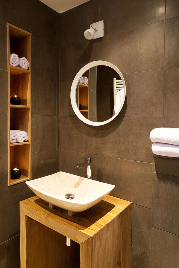 A modern bathroom with a wooden vanity, a round mirror, and a built-in shelf with folded towels.