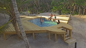 Une terrasse en bois entoure une piscine, avec une plage de sable et des palmiers en arrière-plan.
