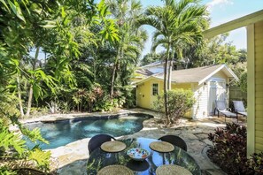 A backyard with a swimming pool, a dining table with chairs, and a small yellow building.