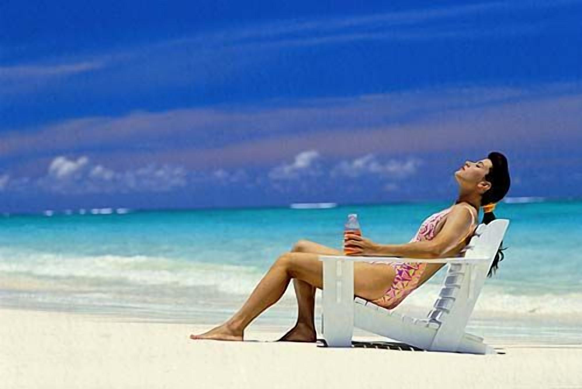 A person reclining on a beach chair with a bottle, overlooking a beach with clear water and a blue sky.