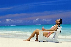 A person reclining on a beach chair with a bottle, overlooking a beach with clear water and a blue sky.