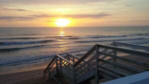 Eine hölzerne Treppe führt zu einem Strand mit Wellen und einem Sonnenuntergang im Hintergrund.