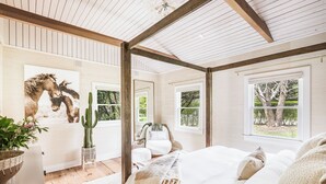 A bedroom with a wooden ceiling, large windows, a bed, a chair, and a cactus.