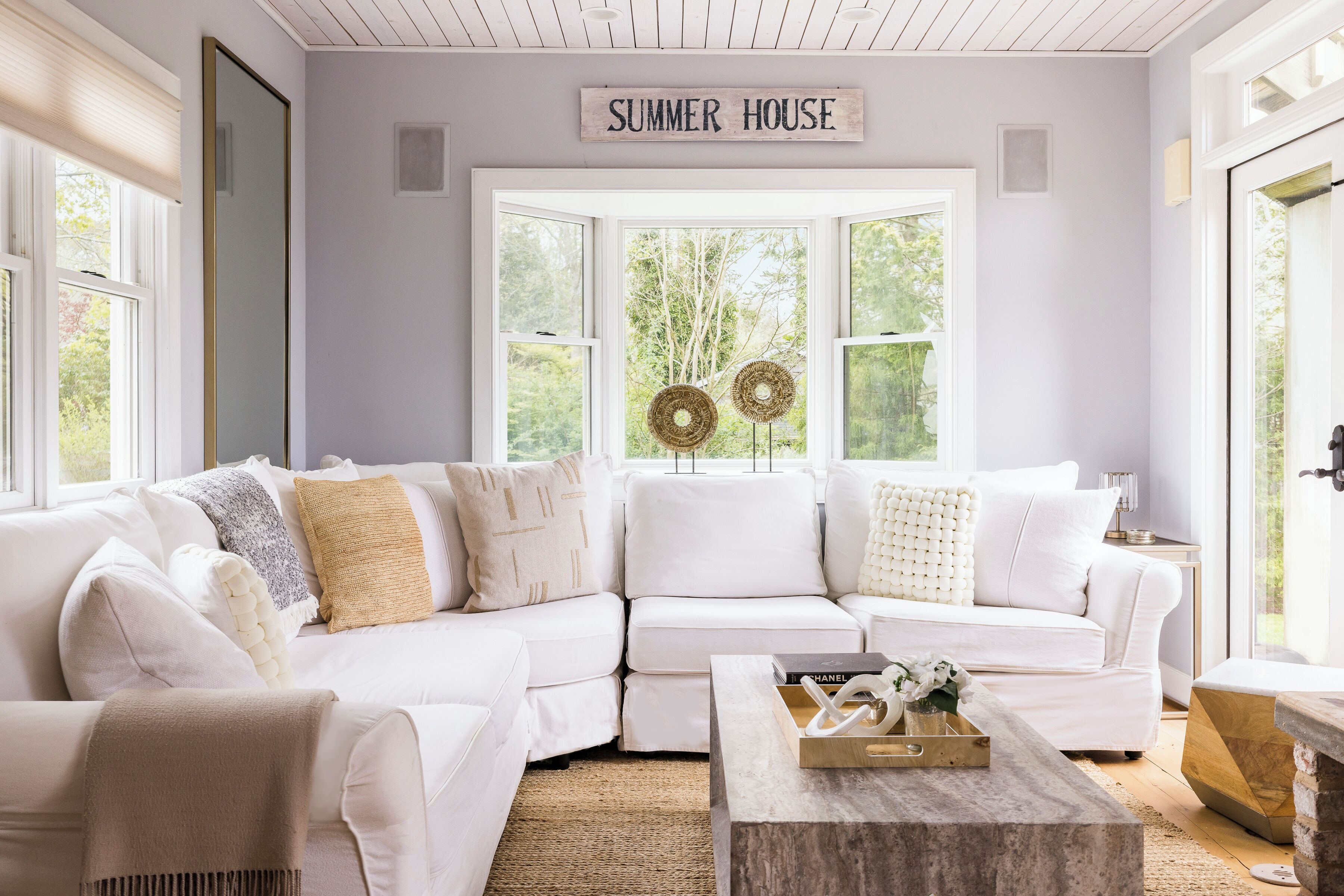 A white sectional sofa with a 'SUMMER HOUSE' sign above a large window, a wooden coffee table, and a woven rug.