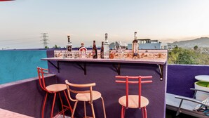 Eine Dachterrasse mit Blick auf Stadt und Berge. Sie bietet eine Bar mit Flaschen und Gläsern sowie drei rote Stühle.