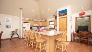 A modern kitchen with wooden cabinets, a central island with bar stools, a dining area with a wooden table and chairs, and a TV in a corner. - 8 BEDROOMS: Book your vacation now! (Corolla)