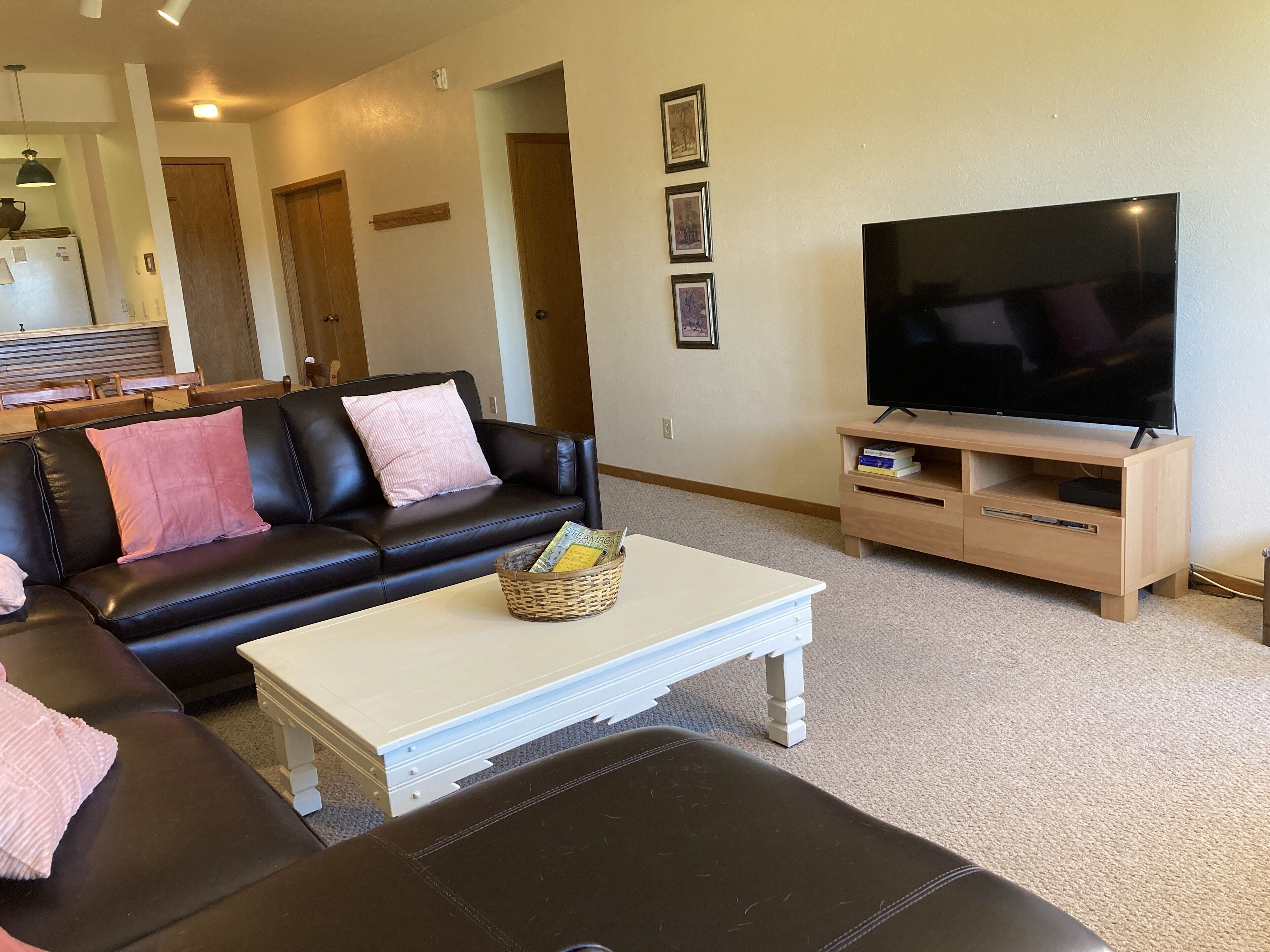 A living room with a black leather sofa, a white coffee table, a TV on a wooden stand, and a kitchen area in the background.