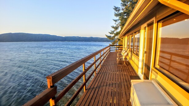 A wooden deck with a railing overlooking a body of water, a row of chairs, and a building with large windows.