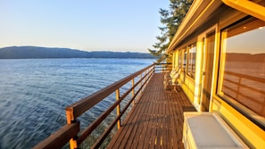 A wooden deck with a railing overlooking a body of water, a row of chairs, and a building with large windows.
