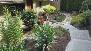 A residential garden with a stone pathway, various plants, and a house with a sloped roof.