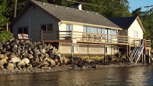 A two-story house with a balcony, situated by a body of water, surrounded by trees.