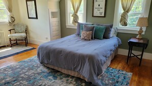Een slaapkamer met een groot bed, een stoel, een dressoir en een raam met gordijnen.