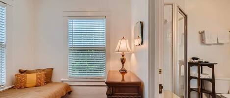 Une chambre à coucher comprenant un lit, une table de chevet, une lampe, une fenêtre avec des stores et une porte donnant sur une salle de bain.