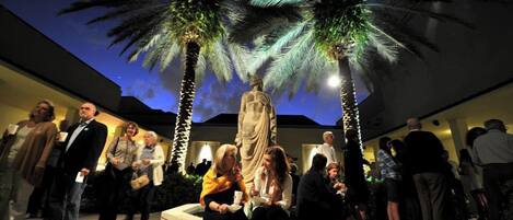 Un groupe de personnes s’est rassemblé autour d’une statue dans un espace extérieur agrémenté de palmiers et illuminé par le ciel nocturne.