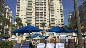 Un hôtel de grande hauteur doté de nombreux balcons, d’un espace piscine avec des chaises longues et des parasols, ainsi que de palmiers.