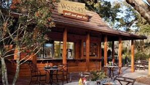 Un espace extérieur aménagé avec une pergola en bois, des tables et des chaises. Une enseigne au-dessus indique : « Woocsy grill et bar au bord de la piscine ».