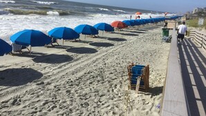 Une plage bordée de rangées de parasols bleus et rouges, une promenade en bois et l’océan en toile de fond.