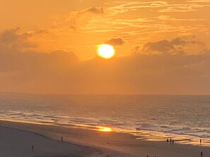A beach scene with the sun setting over the ocean, people walking along the shore, and a clear sky.