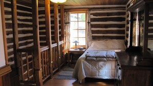 A log cabin room with a bed, wooden walls, a window with curtains, and a lamp on a nightstand.