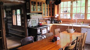 A rustic cabin interior with a wooden dining table, chairs, a stove, and a large window with a view of greenery.