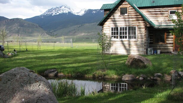 A log cabin with a green roof, a small pond, and mountains in the background.