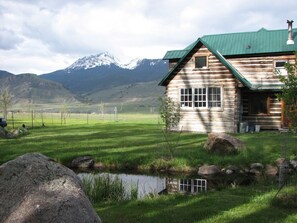 Una cabana de fusta amb un sostre verd, un petit estany i muntanyes al fons.