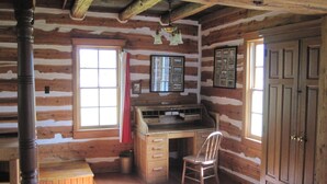 A log cabin interior with a wooden desk, a chair, a window with curtains, and a door.