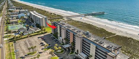 An aerial view of a coastal hotel with a parking lot, a beach, and a pier.