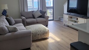 A living room with a sofa, ottoman, and a television on a stand. - Surfside Beach Paradise! Steps to the Beach, Ocean View, Family Friendly (Surfside Beach)