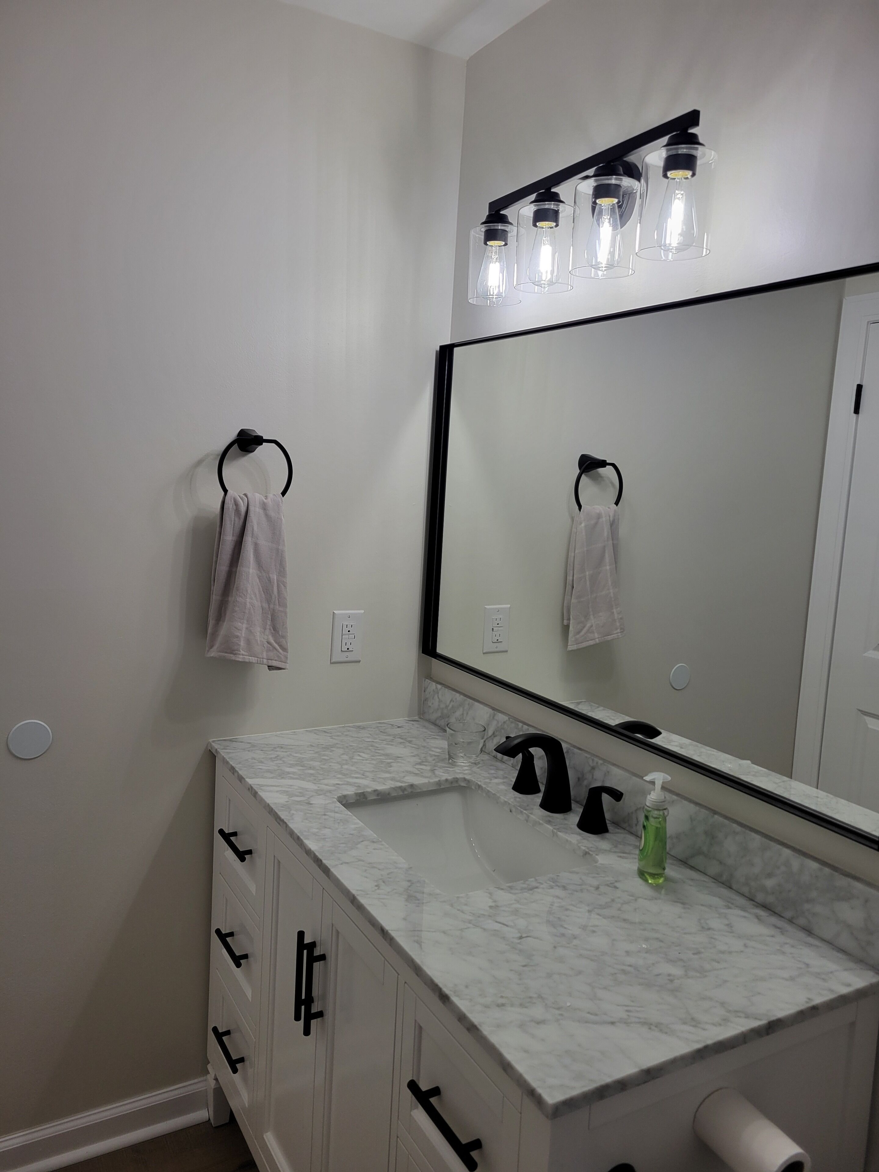 A bathroom with a white marble sink, black faucet, and a large mirror above it. There are two towels hanging on hooks and a green bottle of hand soap on the counter.