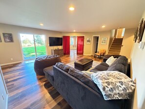 A spacious living room with a large sofa, a coffee table, and a television. There is a sliding glass door leading to a patio area.